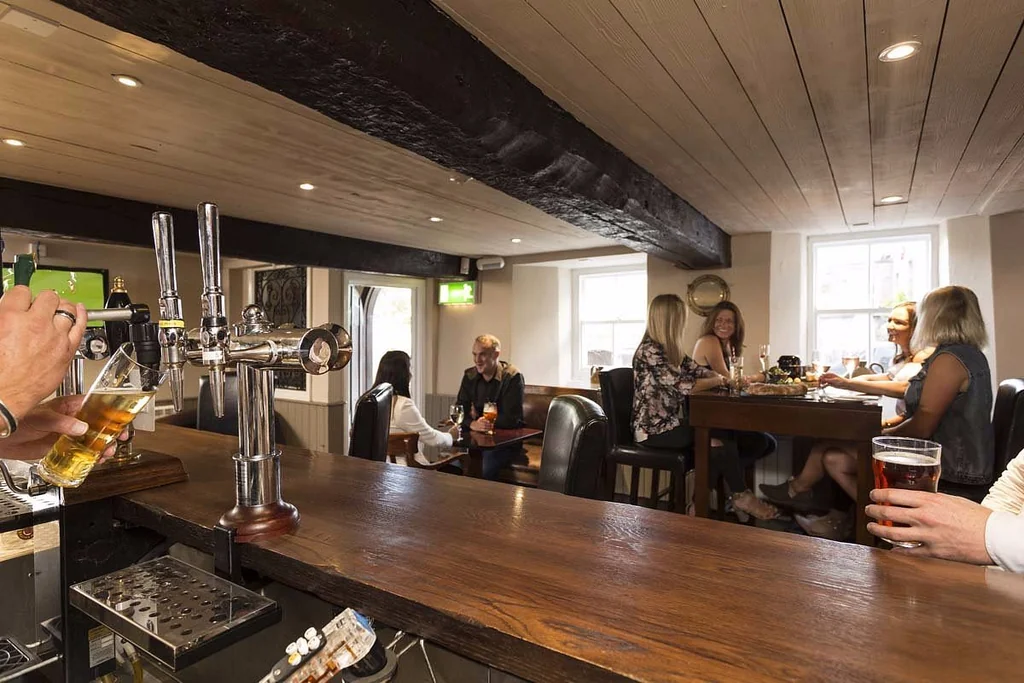 In a cozy bar, a wooden counter holds a tap and a glass of beer, with patrons seated around it. A bartender is nearby, and a television is mounted on the wall. The bar is well-lit, and the atmosphere is relaxed and social.