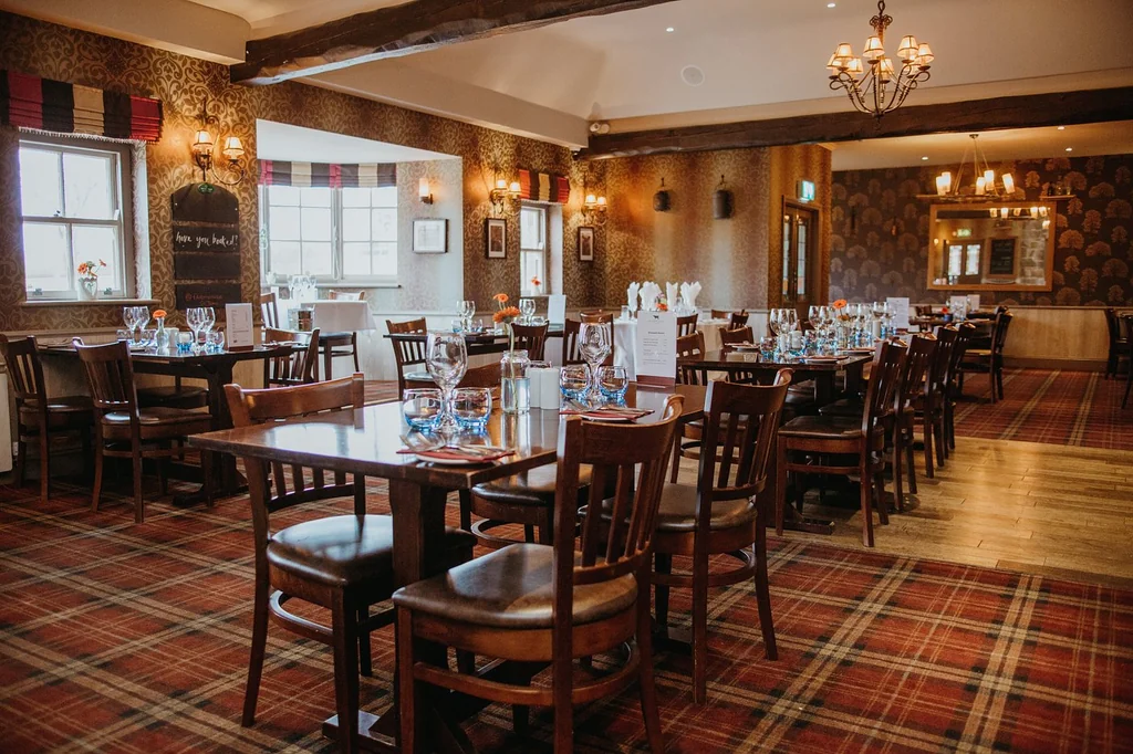 The image shows a cozy, well-lit restaurant with a red and brown checkered carpet, wooden chairs, and tables set for a formal dining experience. The walls are adorned with framed pictures and a chandelier, while the windows offer a view of the outside. The restaurant is ready to welcome guests and offers a warm, inviting atmosphere.