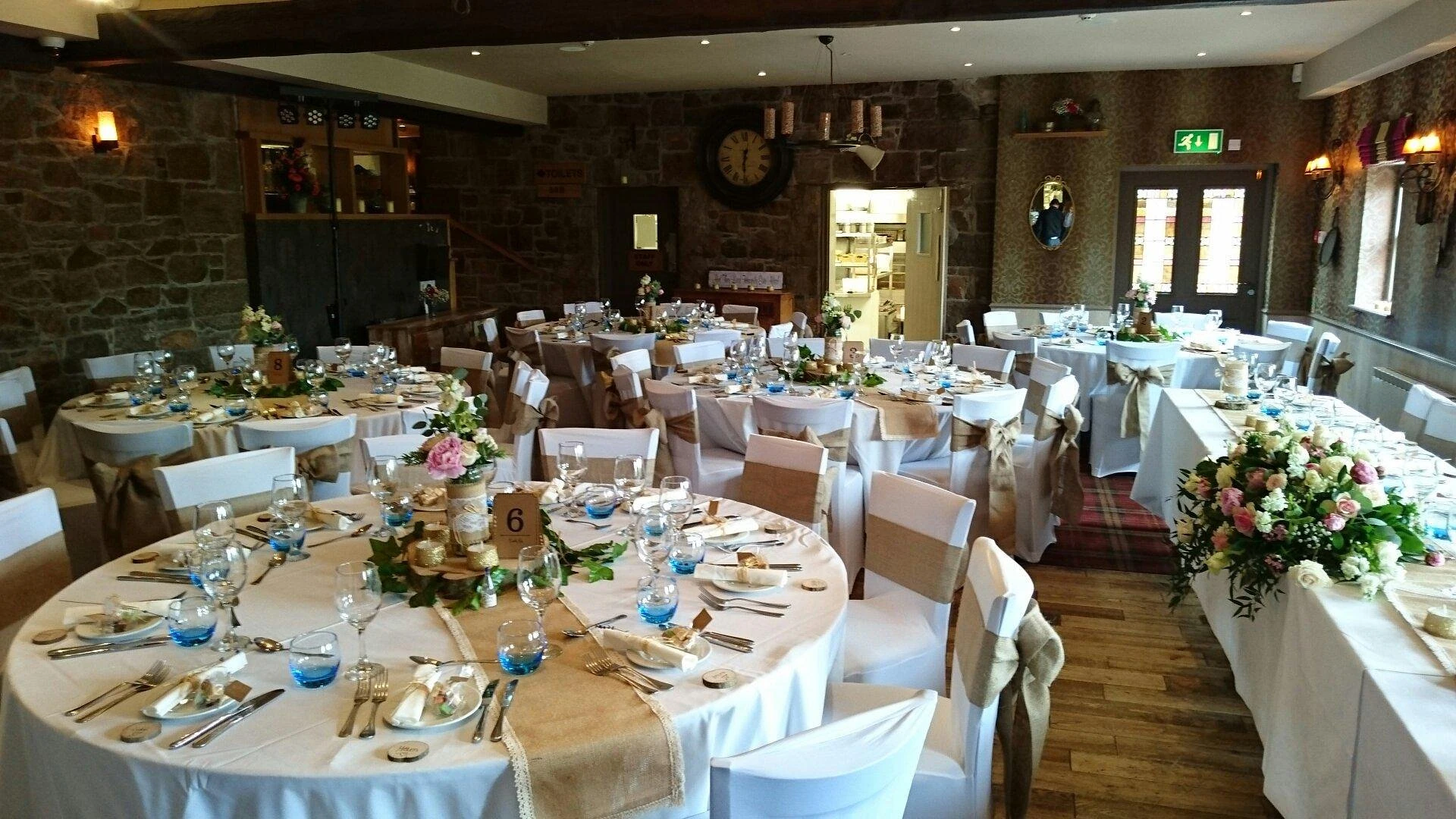 The image shows a beautifully set banquet hall with round tables draped in white tablecloths, adorned with blue and pink flowers. The tables are surrounded by white chairs, and the room features rustic stone walls, a stone fireplace, and a large window. The room is illuminated by natural light and has a warm, inviting atmosphere.