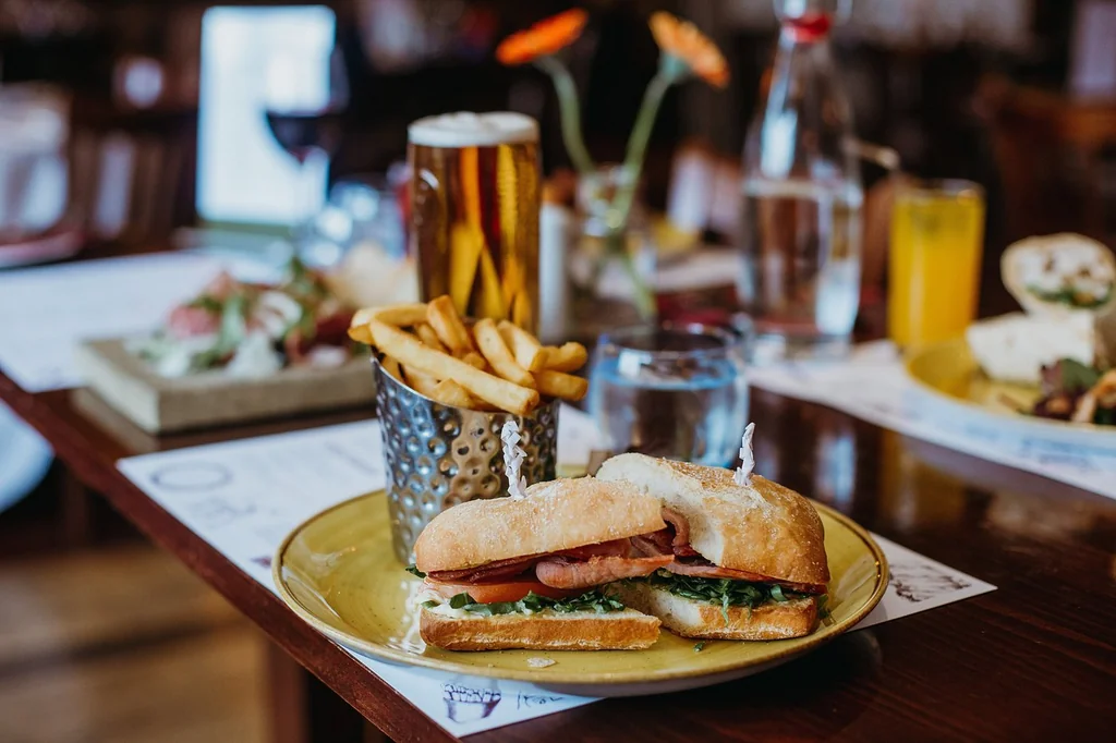 The image features a cozy dining scene with a wooden table, a golden yellow plate with a sandwich and fries, and a glass of water. A vase with orange flowers adds a touch of nature. The arrangement suggests a meal for two, with the sandwich and fries as the main course. The scene is inviting and visually appealing.