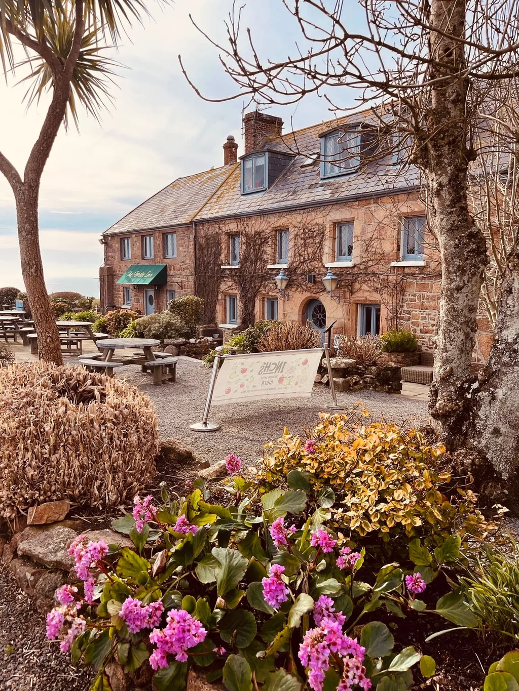 The image shows a serene scene at dusk, with a white building with a red roof and green awning, a red flag, and a garden filled with pink and red flowers. The building is surrounded by a gravel path and picnic tables, inviting visitors to enjoy the surroundings. The sky is a deep orange, suggesting the setting is in the evening. The image captures the tranquility and beauty of the setting.