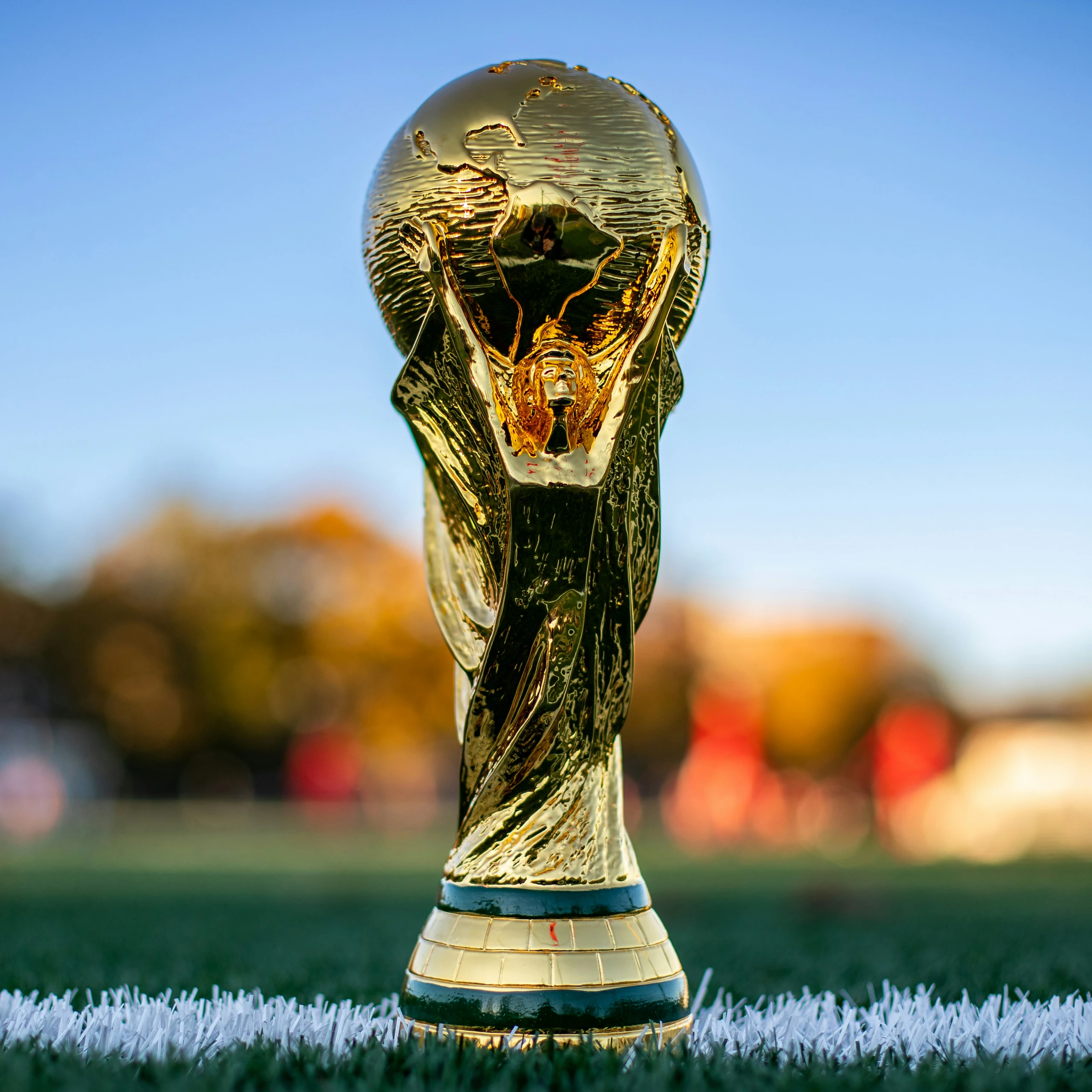 A golden soccer trophy with a human head and wings, adorned with a gold crown, stands on a white soccer field, surrounded by a blurred background of trees and a clear blue sky. The trophy is the central focus, with the field and sky providing context. The image captures a moment of achievement and celebration.