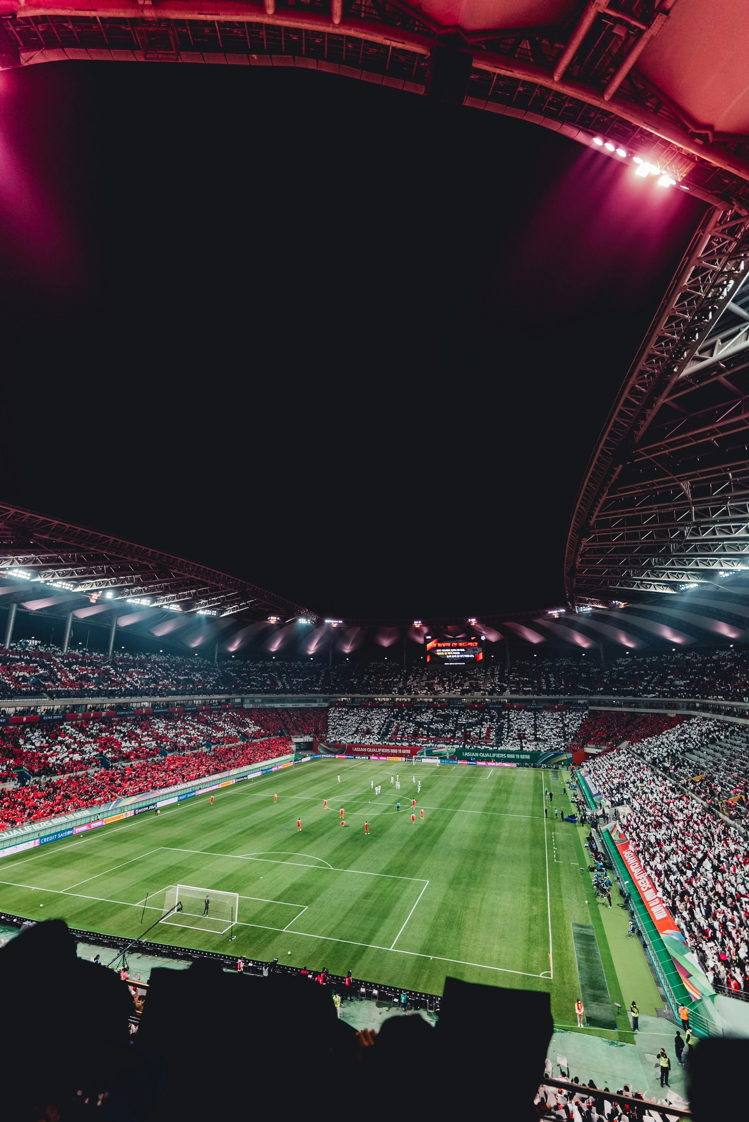 A nighttime soccer match is captured in a large stadium, with a vibrant green field and white boundary lines. Players are scattered across the field, and the stands are filled with excited spectators. The stadium is illuminated by red and white lights, creating a lively atmosphere. The image is taken from the stands, offering a comprehensive view of the game. The landmark "sa_15004" is not identifiable in the image.