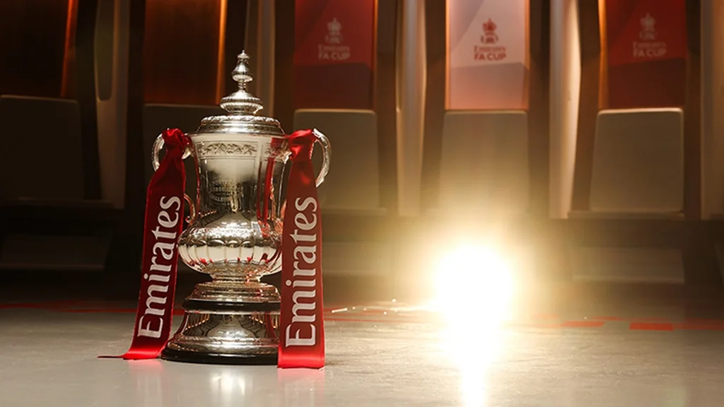 A silver trophy with a pointed top and red ribbon is the central focus of the image, placed on a wooden floor. The trophy is illuminated by a bright light, casting a warm glow on the surrounding area. In the background, red seats and a red banner with the word "LEGO" are visible, suggesting a stadium setting. The image captures a moment of triumph and celebration, with the trophy symbolizing the team's victory.