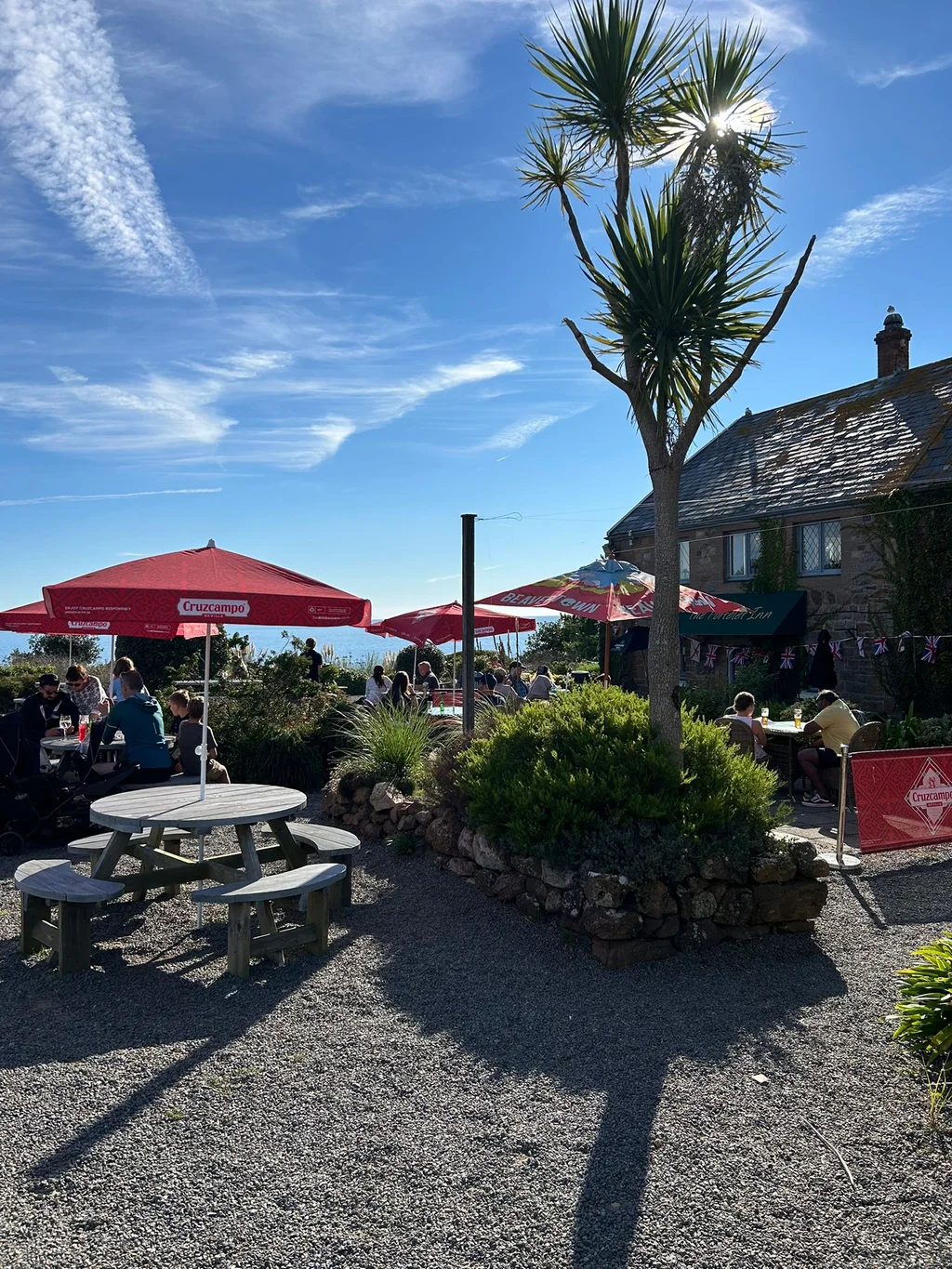 A serene outdoor café on a rocky beach is the focal point of the image, with a large red umbrella providing shade. The café is surrounded by a stone wall and a palm tree, while people enjoy the view. The clear blue sky and ocean add to the tranquil atmosphere.