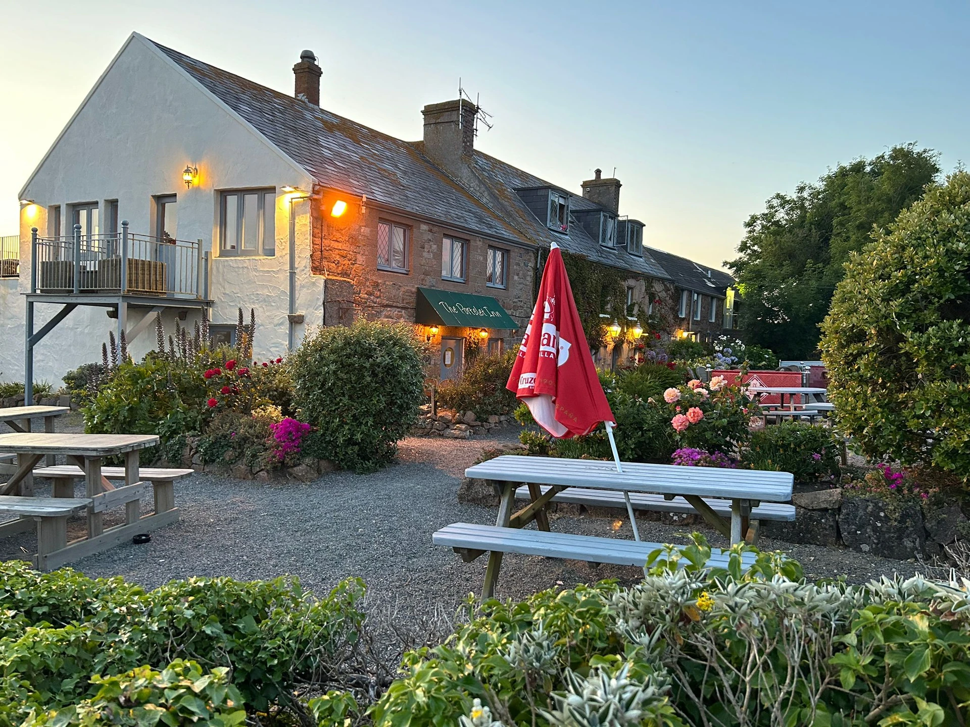 The image shows a serene scene at dusk, with a white building with a red roof and green awning, a red flag, and a garden filled with pink and red flowers. The building is surrounded by a gravel path and picnic tables, inviting visitors to enjoy the surroundings. The sky is a deep orange, suggesting the setting is in the evening. The image captures the tranquility and beauty of the setting.