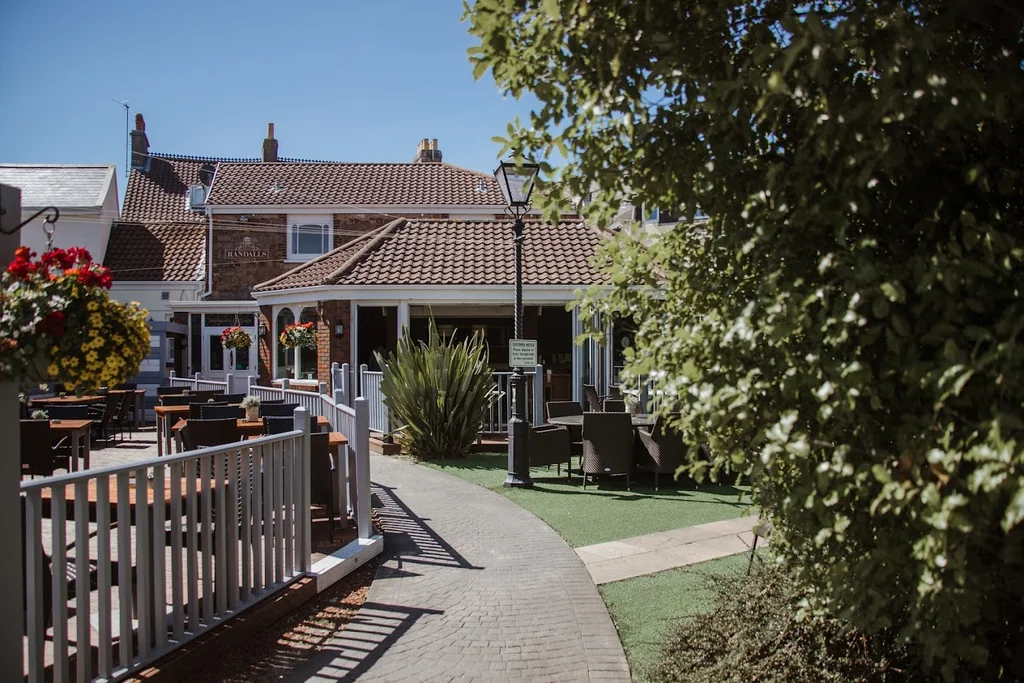 A serene residential area features a white fence, a white picket fence, and a patio with tables and chairs. A red-roofed building with a white roof is in the background, and a lamppost stands nearby. The scene is set against a clear blue sky, creating a tranquil and inviting atmosphere.