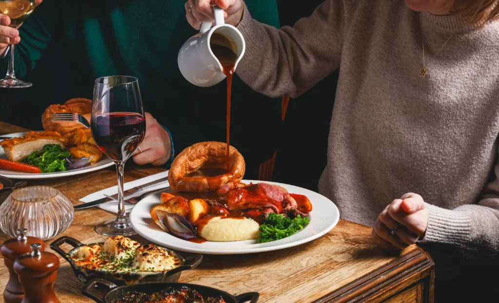A cozy dining scene features a wooden table with various dishes, including a large plate of meat, a plate of potatoes, a plate of green salad, and a plate of bread. Two people are present, one pouring red sauce over the meat and the other holding a glass of wine. A salt shaker, pepper shaker, and a black pepper grinder are also present. The table is adorned with a silver fork, a white plate, and a white bowl. The scene is warm and inviting, with a sense of anticipation for the meal.