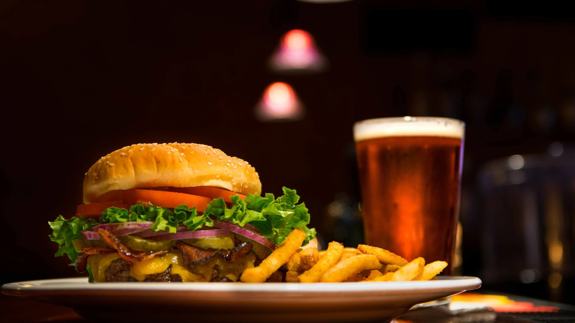 A burger with a bun, lettuce, tomato, and onion is on a white plate, accompanied by a side of fries. A glass of beer is to the right, with a red lamp in the background. The scene is set in a dimly lit bar or restaurant.