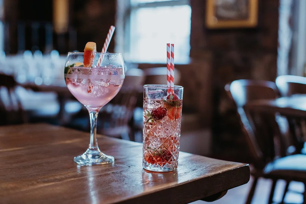 In a cozy restaurant, two glasses of pink and red drinks sit on a wooden table, each with a straw and a lemon slice. The pink glass has a red strawberry, while the red glass has a strawberry and a lime slice. The background features wooden chairs and a window, creating a warm and inviting atmosphere.