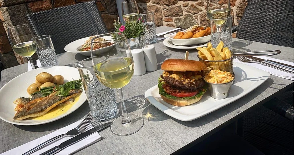 A dining table is set with a variety of food and drink items, including a large burger, fries, potatoes, and a salad. The table is adorned with silverware and wine glasses, and a vase with a flower adds a touch of nature. The scene is set against a rustic stone wall, creating a cozy and inviting atmosphere.