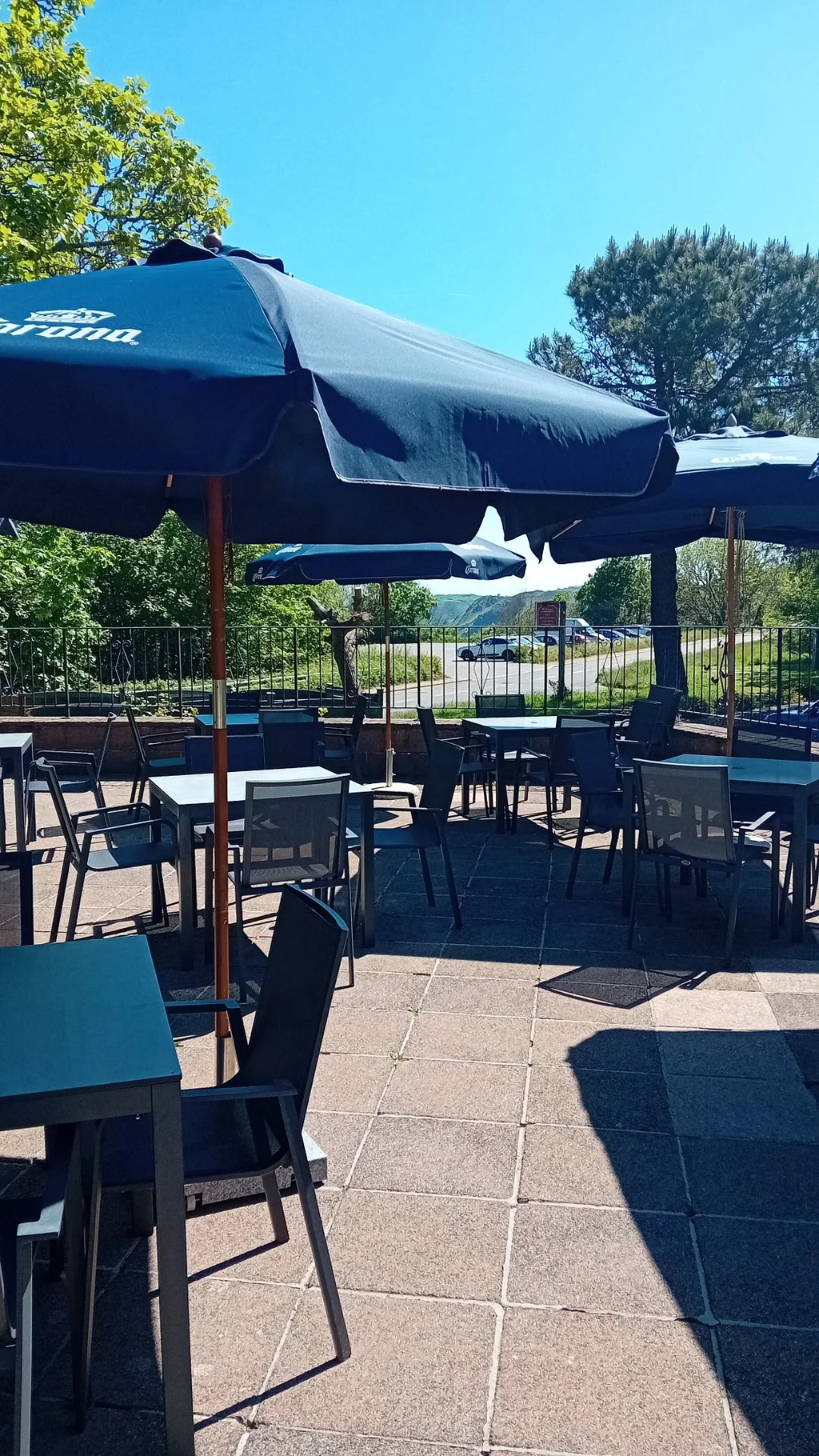 The image shows a serene outdoor patio area with six tables and chairs under two large blue umbrellas. The tables are arranged in a U-shape, and the chairs are black. The patio is surrounded by a fence and trees, with a clear blue sky above. The scene is peaceful and inviting, perfect for a quiet afternoon.