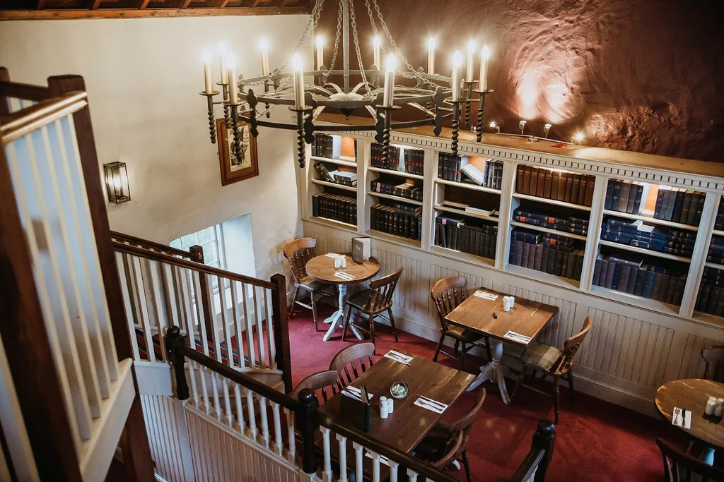 The image shows a cozy, well-lit restaurant with wooden tables and chairs, a bookshelf filled with books, and a chandelier casting a warm glow. The red carpet adds warmth, and a staircase leads to the second floor. The scene is inviting and comfortable, perfect for a meal or a quiet evening.