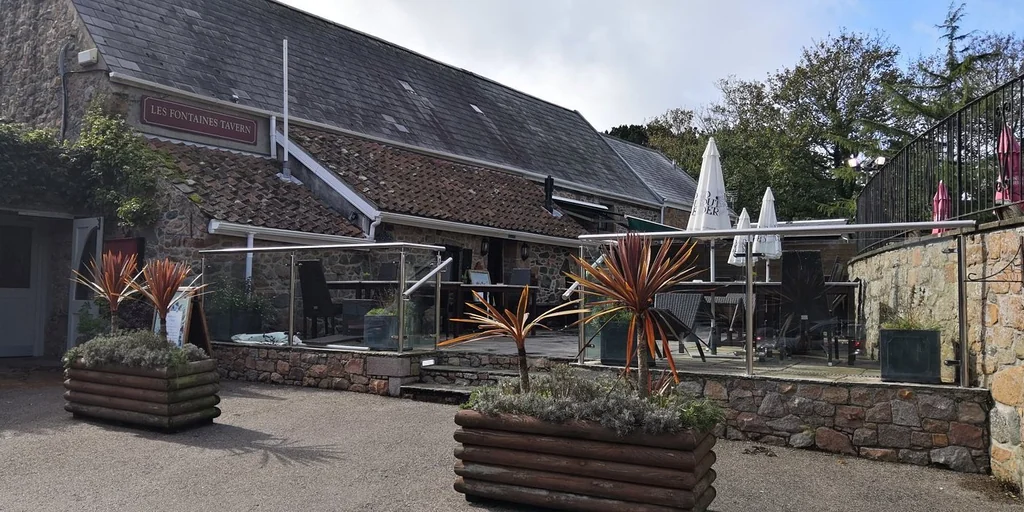 The image shows the historic "The Old Inn" pub in the UK, with a black tiled roof and white awning. The pub is surrounded by a stone wall and a stone fence, and features a wooden table and chairs. The scene is set against a backdrop of trees and a clear blue sky, creating a serene atmosphere.