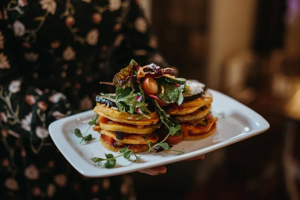 A person in a floral dress holds a white plate with golden brown pancakes, garnished with fresh greens and a red sauce, against a blurred background.