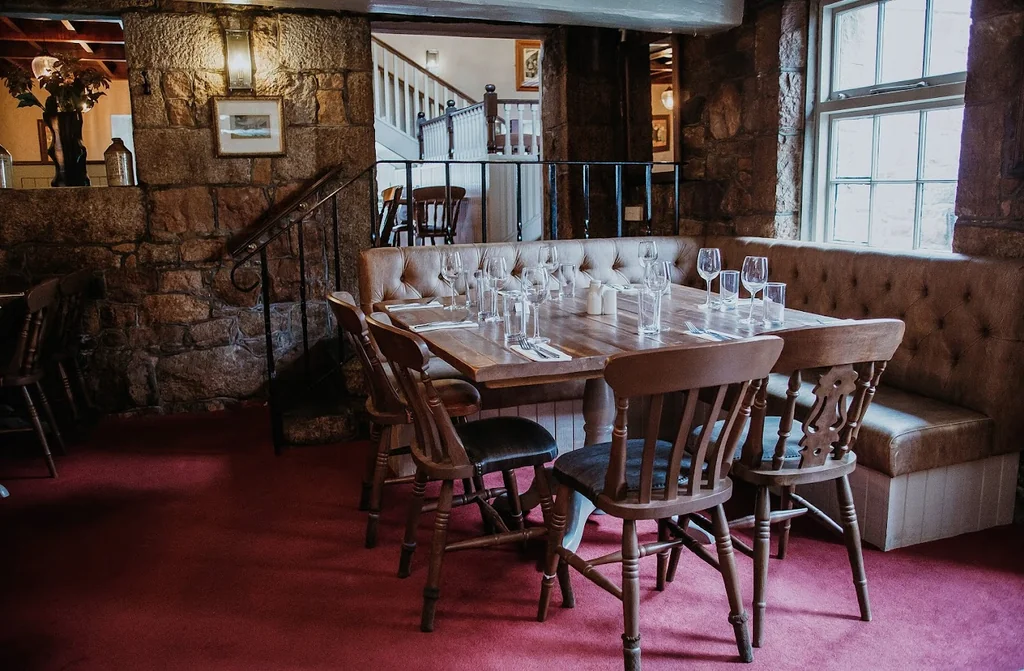 A cozy restaurant interior features a long wooden table with white tablecloths and silverware, surrounded by wooden chairs with leather backs. The table is set for a meal, with wine glasses and silverware. The walls are adorned with paintings and a chandelier, while a bench and a window provide seating. The floor is covered in a red carpet, and a white sofa is present. The room is well-lit and has a rustic charm.