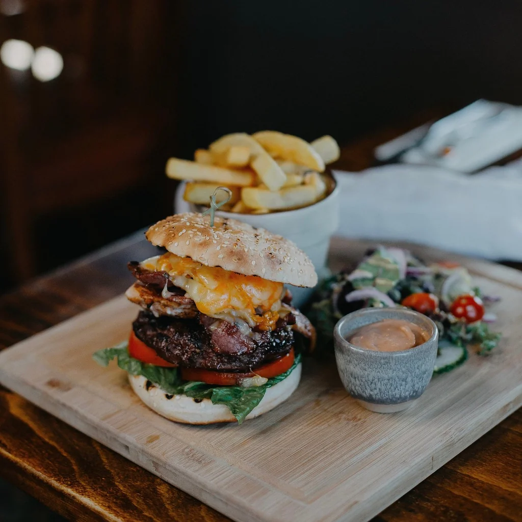 A wooden cutting board holds a delicious burger with a bun, cheese, tomato, lettuce, and pickles, accompanied by golden fries and a small bowl of sauce. The scene is set on a wooden table, with a knife and fork nearby.
