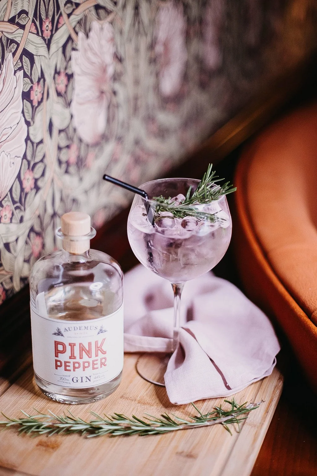 A wooden table holds a bottle of "Pine & Rose" vodka, a cocktail glass with a sprig of rosemary, and a pink napkin. The table is near a couch and a floral wallpaper.