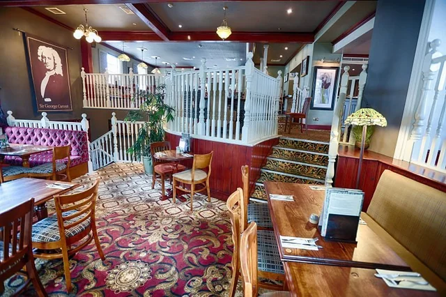 The image depicts a spacious, well-lit restaurant with a warm, inviting atmosphere. The room features a variety of wooden furniture, including tables, chairs, and benches, with a red and gold carpet covering the floor. The walls are adorned with pictures and a white staircase, while hanging lights and a chandelier illuminate the space. A large plant adds a touch of nature, and a white railing provides safety. The restaurant is ready for guests to enjoy a meal and conversation.