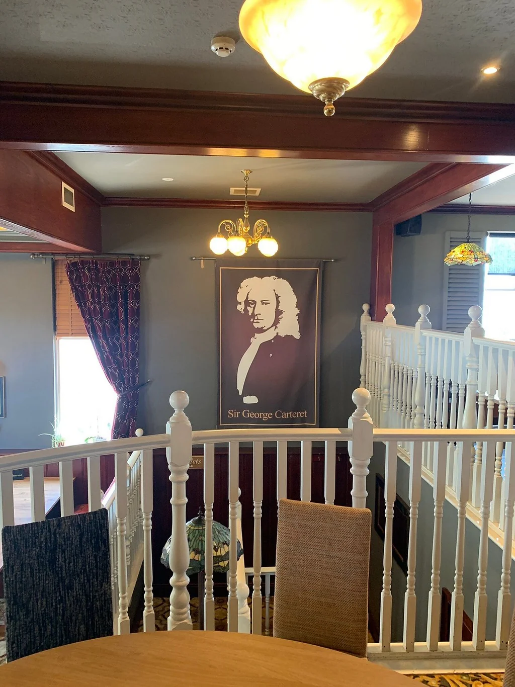 A cozy room features a wooden floor, white railing, and a wooden ceiling. A framed portrait of a man with glasses and a black and white photograph of a man with glasses are on the railing. A yellow chandelier hangs above, casting a warm glow. A wooden table with a vase and a black and white photograph are on the floor, and a red curtain adds color. The room is inviting and well-organized.