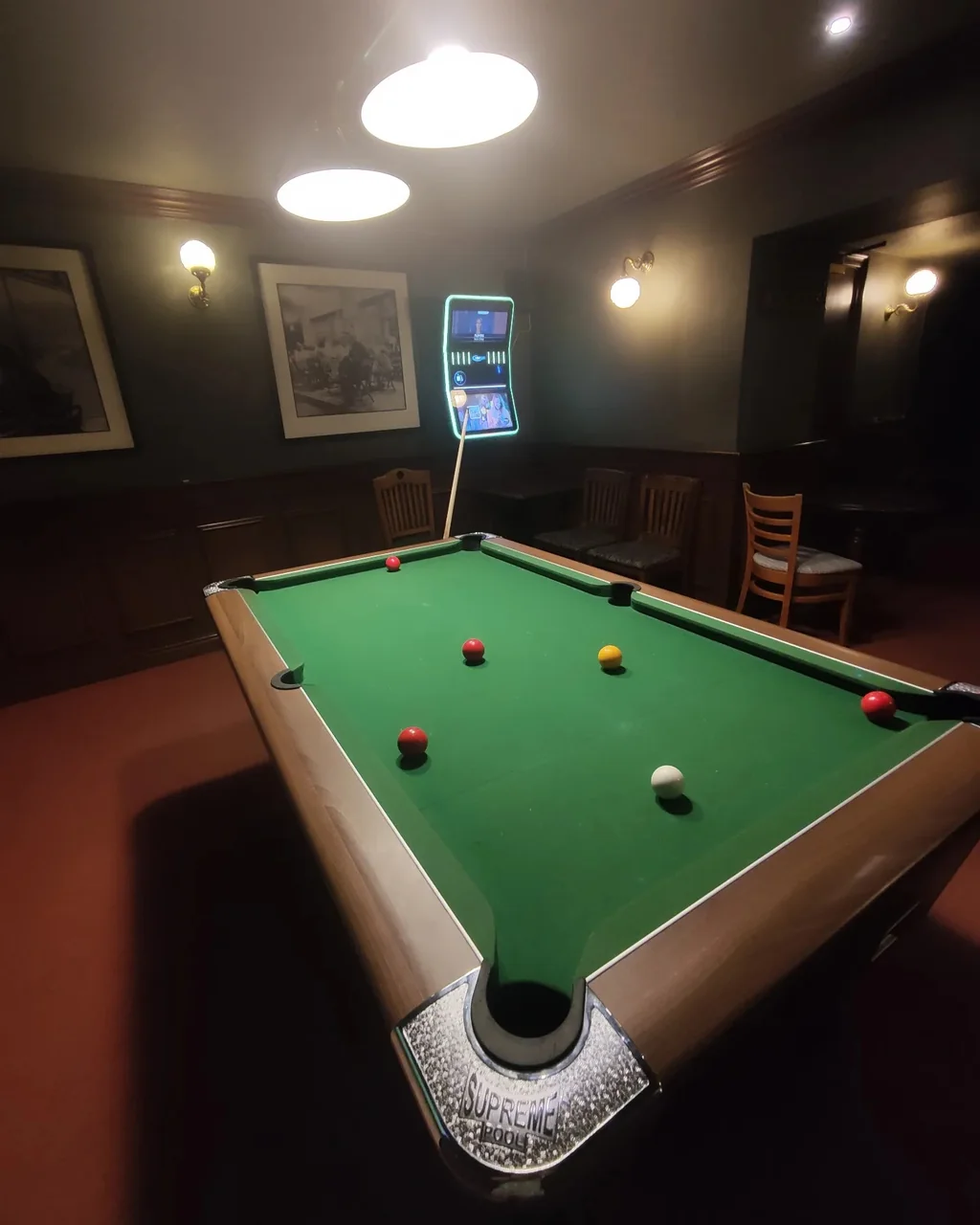 A dimly lit billiards room features a green pool table with a white net, surrounded by colorful pool balls. A phone is on the table, and a wooden chair is in the background. The room has dark wood paneling and framed pictures on the walls. The scene is serene and inviting, with the billiards table as the central focus.
