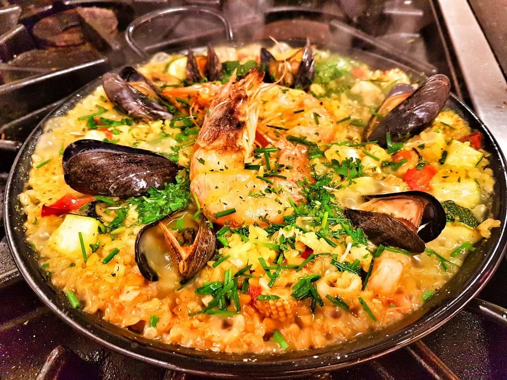 A large skillet filled with a vibrant seafood and rice dish is on a stovetop, with a black pan beneath it. The dish features shrimp, clams, mussels, and vegetables, and is seasoned with herbs. The stovetop is in the background, with a black stove and burners visible.