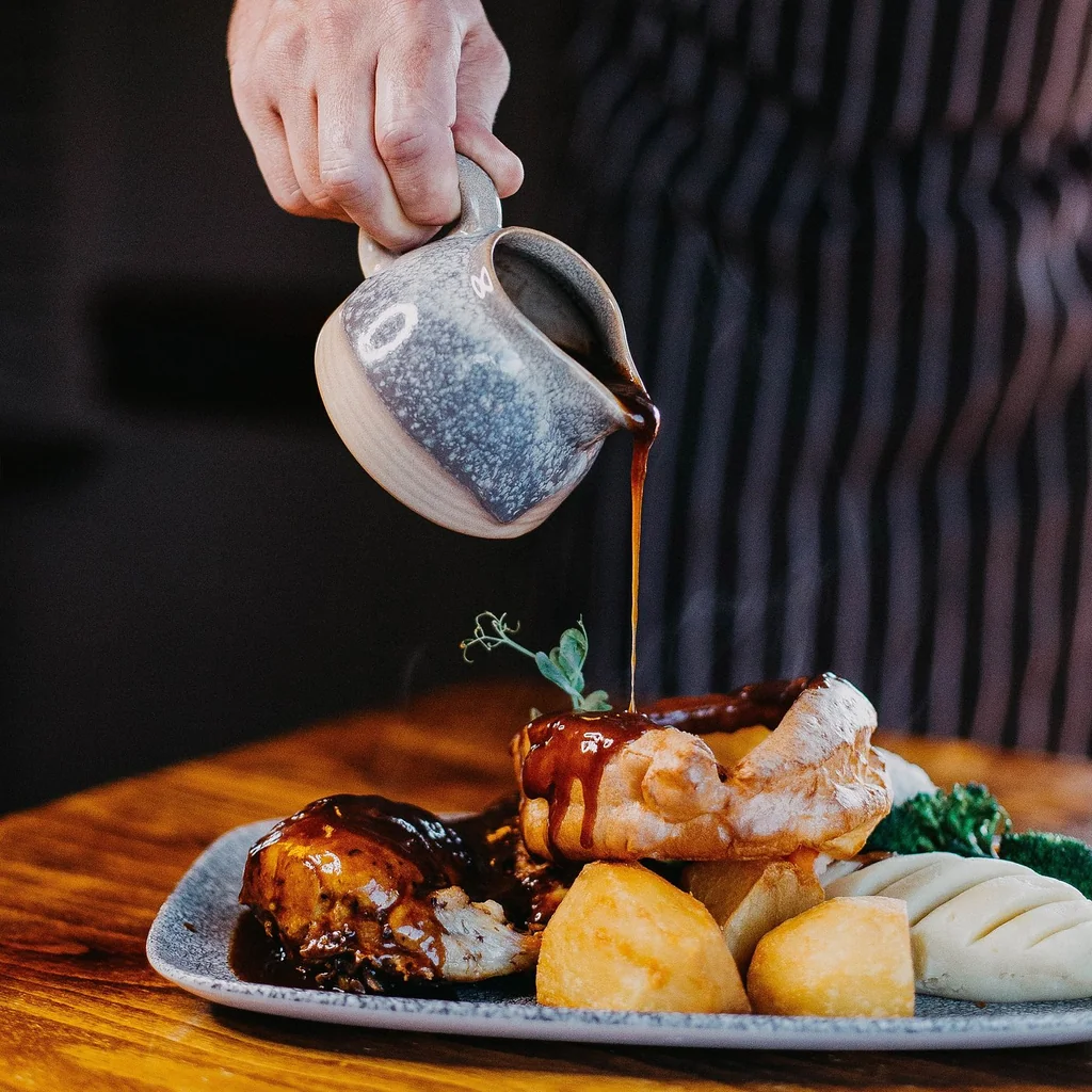 A person in a black and white striped apron is pouring gravy over a plate of food on a wooden table, with a white plate and a silver pitcher. The plate features a meat dish, bread, potatoes, and vegetables. The background is blurred, emphasizing the main subject and the food.