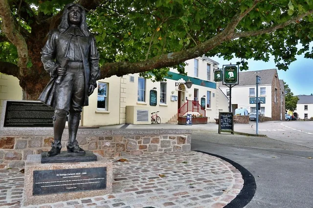 A bronze statue of a man in a suit and hat stands on a cobblestone street, with a tree and a building in the background. The statue is on a pedestal, and the scene is framed by a tree and a building. The image is taken from a low angle, emphasizing the statue's prominence.