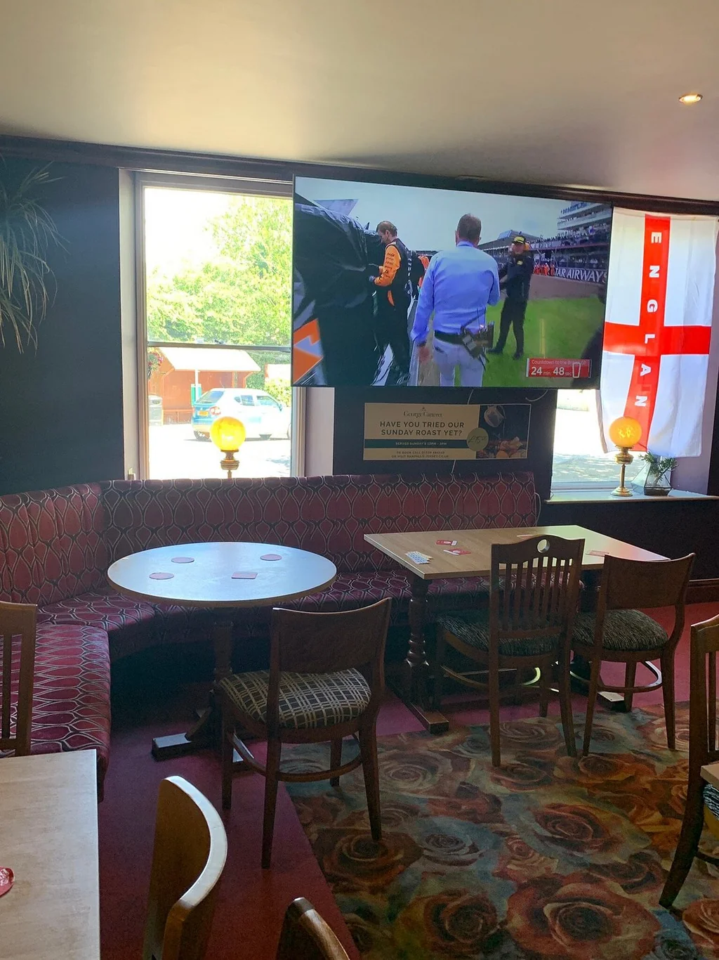 The image shows a cozy, modern restaurant with a red and black patterned carpet, a wooden table with a white tablecloth, and a red sofa with a blue and white striped cushion. The walls are adorned with posters of a soccer game and a football match, and a large window offers a view of a street. The restaurant is well-lit and inviting, with a warm and inviting atmosphere.