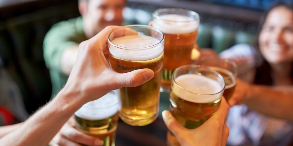 Four hands raise beer glasses in a lively bar setting, with the central person holding a glass filled with golden beer. The background is blurred, focusing on the group and their joyous celebration.