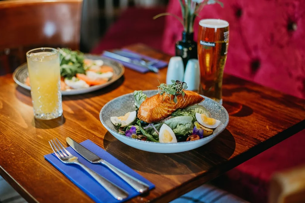 A wooden table in a restaurant setting features a salmon dish with lemon wedges, a salad, and a glass of beer. A blue napkin and silverware are on the table, with a red cushion in the background. The scene is inviting and well-prepared for a meal.