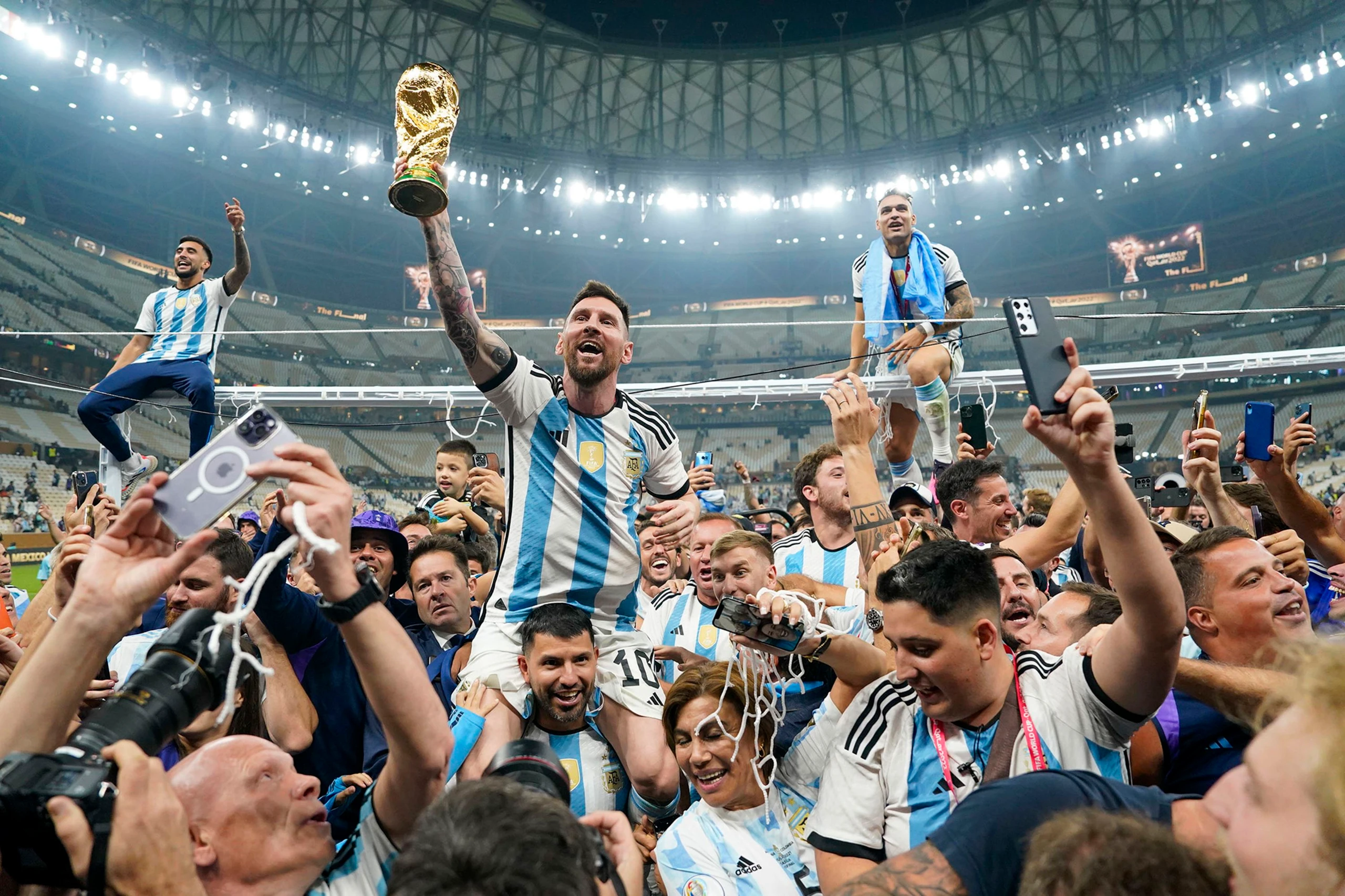 A jubilant soccer player in a blue and white striped jersey holds a gold trophy above his head, surrounded by a sea of fans in blue and white striped jerseys. The stadium is filled with spectators, some holding phones, capturing the moment. The atmosphere is celebratory and triumphant, with the player's victory symbolizing a new chapter in his career.