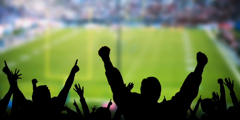 A soccer match is captured in a vibrant, green field with white lines. The crowd, silhouetted, raises their arms in celebration, with some raised to the sky. The image is taken from a low angle, emphasizing the height of the crowd and the stadium's grandeur. The colors are predominantly green, blue, and white, creating a lively and energetic atmosphere.