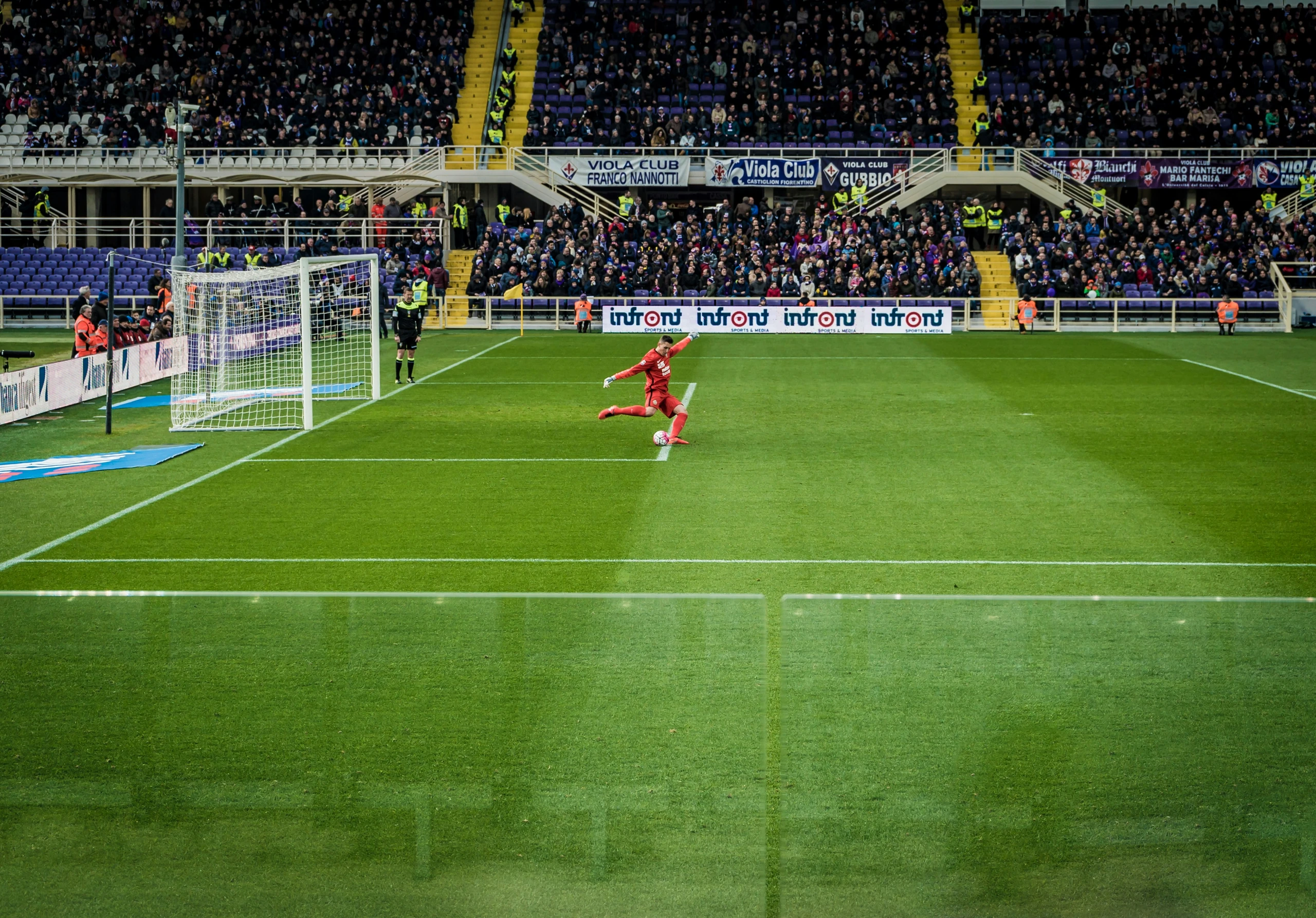 A soccer match is captured in a vibrant stadium, with a player in a red jersey kicking the ball towards the goal. The goal is visible in the background, and the stands are filled with spectators. The image is taken from the side of the field, offering a comprehensive view of the action. The scene is filled with energy and excitement, showcasing the sport's beauty and intensity.