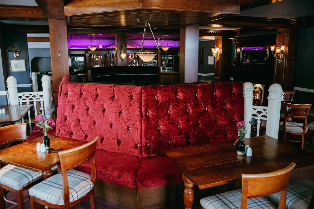 A luxurious bar or restaurant features a large red velvet sofa with a tufted backrest, surrounded by white and gray striped chairs. The space is illuminated by chandeliers and a large window, with a bar and a pool in the background. The atmosphere is cozy and inviting, with a mix of modern and traditional elements.