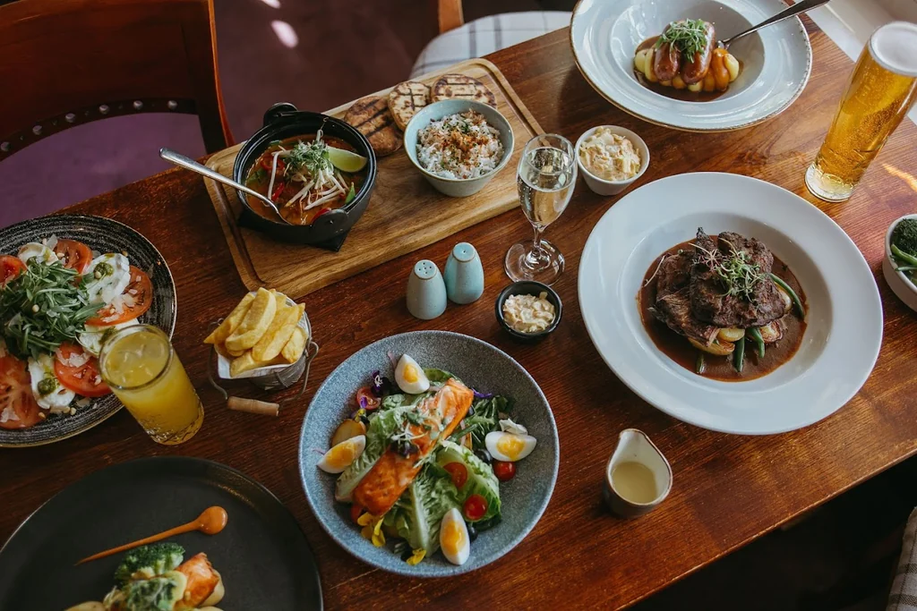 A wooden table is set with a variety of dishes, including a large steak, salad, fries, and salad, accompanied by drinks and condiments. The table is surrounded by a purple chair and a wooden tray, creating a cozy and inviting atmosphere.