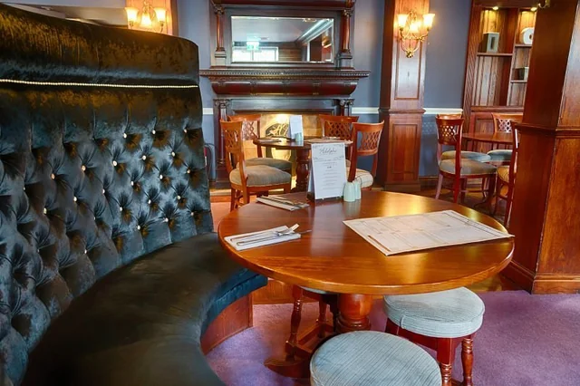 A cozy, vintage-themed bar or restaurant features a round wooden table with a menu, a black velvet booth, and a wooden bar with a mirror. The room has a warm, inviting atmosphere with a purple carpet, wooden furniture, and a fireplace. The bar is illuminated by a chandelier and a mirror, and the overall design is elegant and sophisticated.