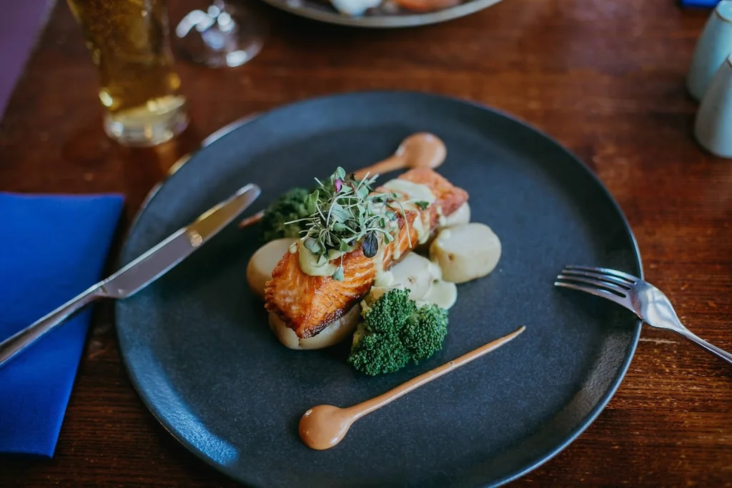 A blue plate with a salmon dish, garnished with a green herb and white sauce, is the centerpiece of a dining scene. The plate is on a wooden table, with a blue napkin and fork nearby. A glass of beer and a glass of water are in the background, creating a relaxed atmosphere. The image captures a moment of anticipation before a meal.