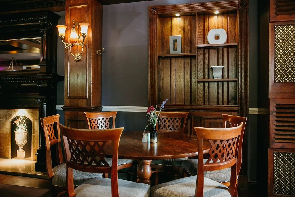 A cozy dining room features a round wooden table with four chairs, surrounded by a wooden bookshelf and a fireplace. A chandelier casts a warm glow, and a vase with a flower adds a touch of nature. The room is illuminated by a lamp, creating a warm and inviting atmosphere.