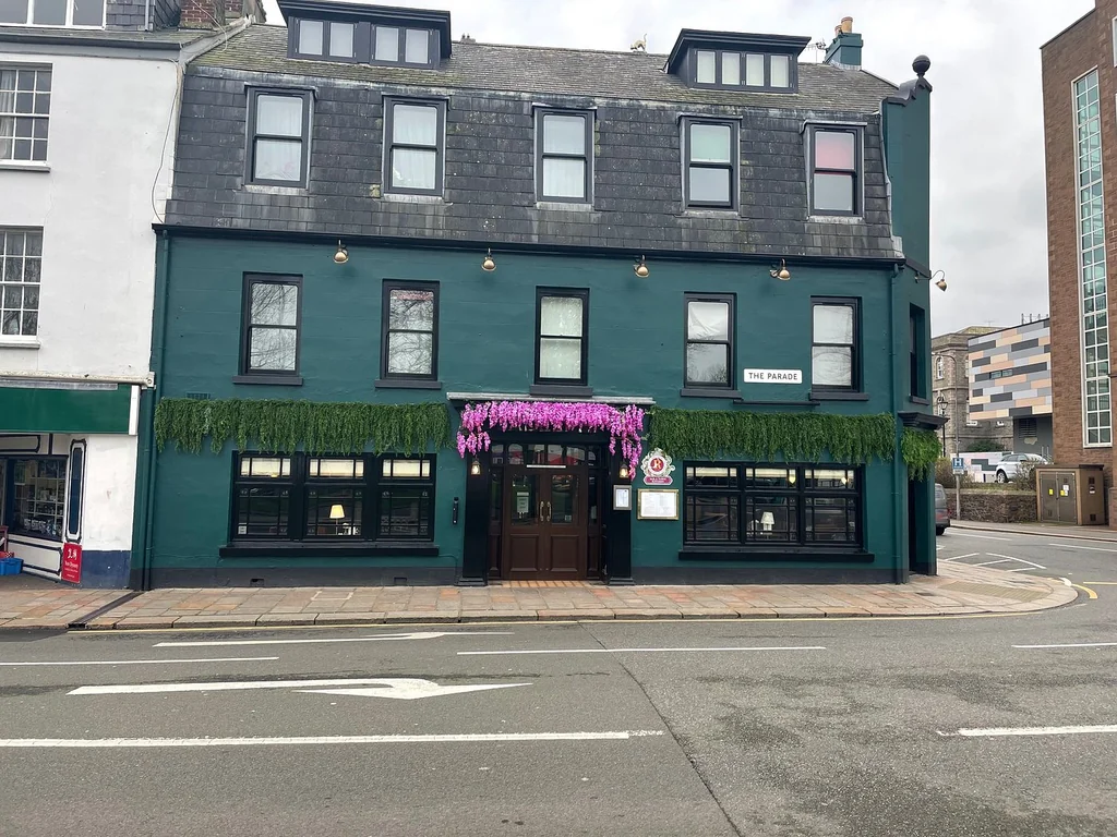 A two-story green building with a black roof and white trim is located on a street corner, featuring a black awning with a pink ribbon and a white sign. The building has six windows with green plants and a red fire hydrant. The street is lined with other buildings, and the sky is overcast. The scene is peaceful and inviting.