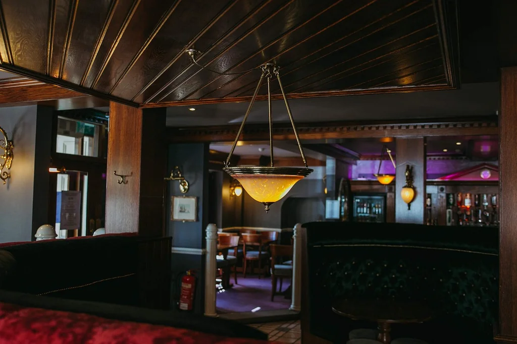 The image shows a cozy bar or restaurant with a wooden ceiling, a hanging lamp, and a red velvet booth. The lamp casts a warm glow, and the room is illuminated by a red fire extinguisher. The background features a mirror, a bar, and a door, creating a warm and inviting atmosphere.