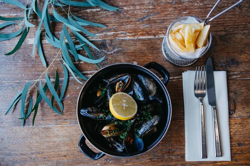 A wooden table features a black frying pan filled with mussels and a lemon slice, accompanied by a white napkin, fork, knife, and a glass of fries. A green leaf adds a touch of nature. The scene is set for a meal, inviting the viewer to enjoy the meal.