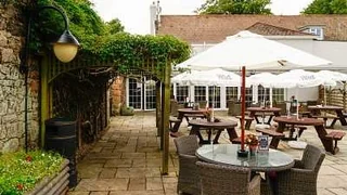 A serene outdoor setting features a stone patio with wicker chairs and tables, surrounded by a wooden fence and a vine-covered wall. A large white umbrella provides shade, and a black trash can is nearby. The patio is bordered by a wooden fence and a brick wall, with a white building in the background. The scene is filled with natural elements and a sense of tranquility.