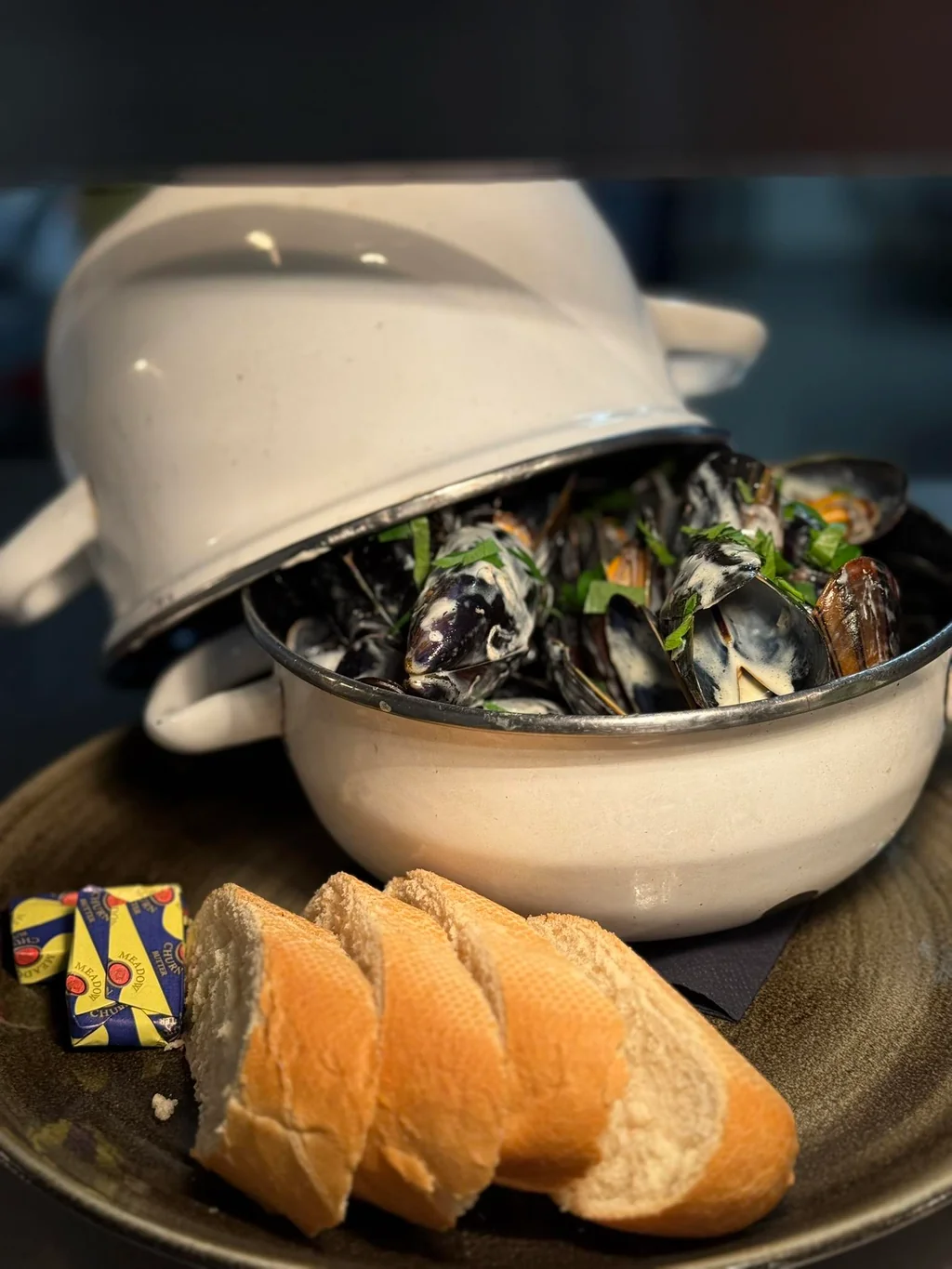 A white crockpot filled with mussels and clams sits on a black plate, with three slices of bread nearby. A colorful bag of crumbs is on the plate, and the crockpot is on a gray tablecloth. The scene suggests a meal in progress, with the crockpot and bread ready to be served.