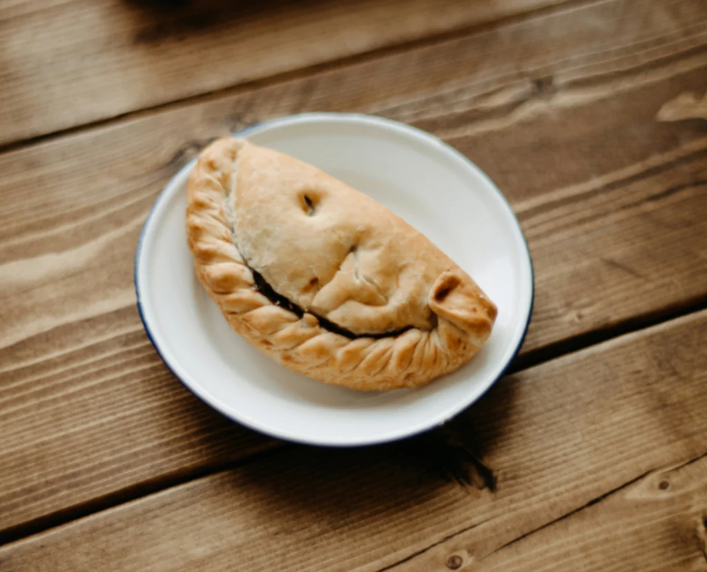 A golden-brown pie slice with a crumpled crust sits on a white plate with a blue rim, resting on a wooden table. The background is blurred, focusing on the pie and plate.