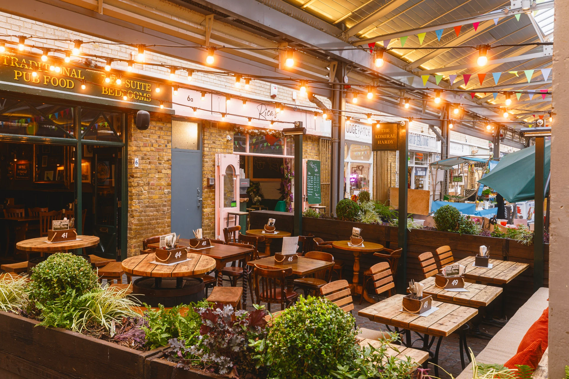 The image shows a lively outdoor restaurant patio with wooden tables and chairs, adorned with white plates and silverware. The patio is surrounded by plants and a brick wall, with a green awning providing shade. The restaurant name "The Kitchen" is displayed above the awning. The perspective is from the ground, looking up at the patio, creating a sense of depth and inviting passersby to enjoy the ambiance.