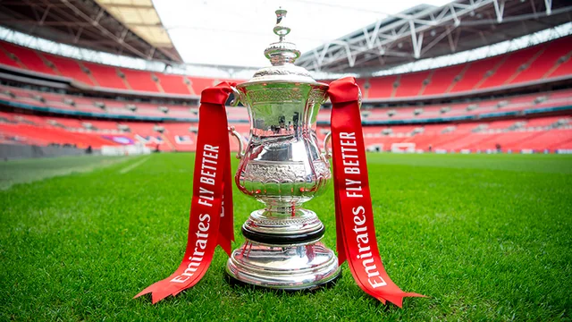 A silver trophy with a white base and red ribbon is prominently displayed on a green field, surrounded by empty red stadium seats. The trophy's position and the background suggest a significant achievement, possibly a World Cup or other major tournament.