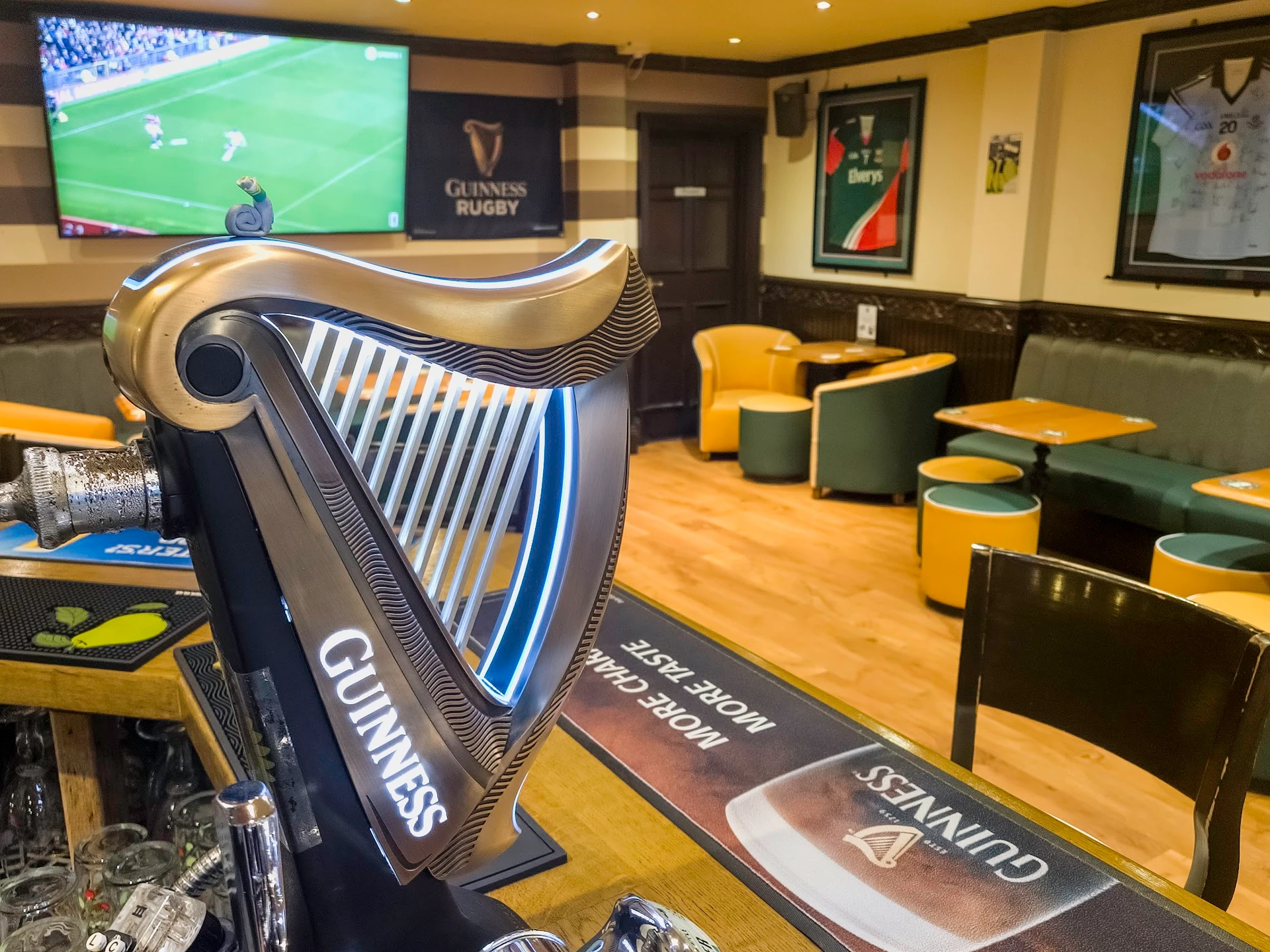 In a英超 stadium, a black and gold St. John's harp stands on a wooden bar, with a "St. John's" sign above. A TV screen displays a match between Manchester United and Arsenal. Green and yellow bar stools and a "St. John's" logo are in the background. The scene is warm and inviting, with a sense of anticipation for the football match.