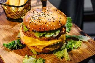 A golden-brown burger with sesame seeds, melted cheese, and lettuce sits on a wooden cutting board atop a black table. A yellow French fries basket is nearby, and a black basket with a wire is in the background. The scene is set against a black table, creating a visually appealing and appetizing meal.