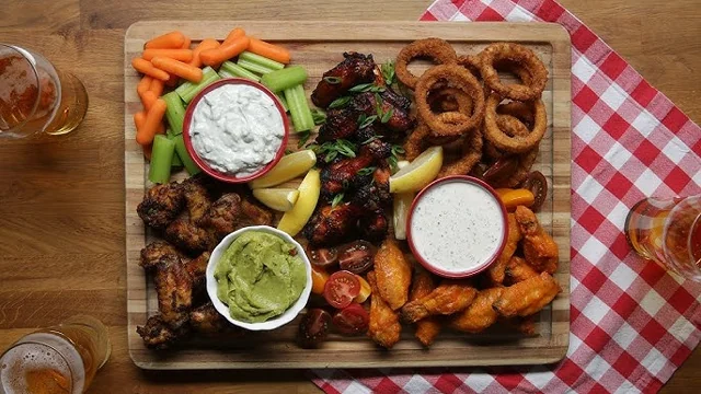 A wooden tray on a red and white checkered tablecloth holds a colorful assortment of wings, chicken wings, caramelized onions, pickles, and guacamole, with two glasses of beer nearby.